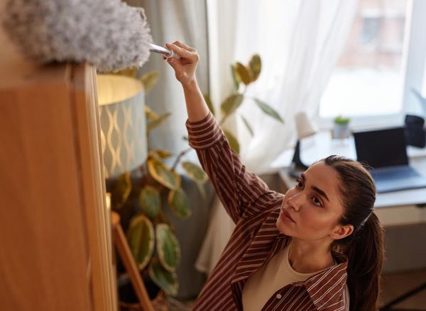 A woman dusting up high on top of a book shelf