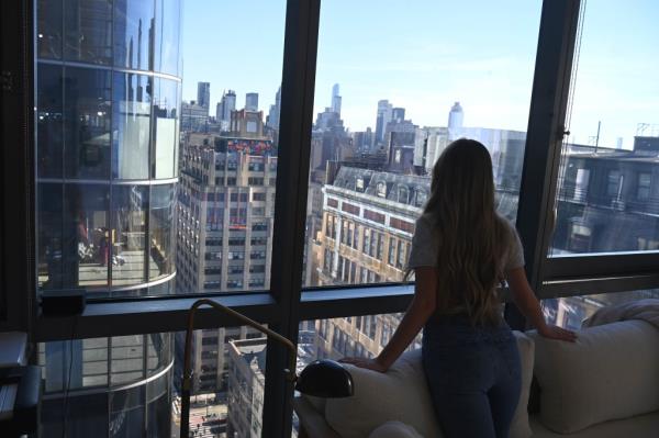 Lilyan seen from behind peering out windows in her apartment, which look out on Two Manhattan West and Kaufman Hall